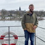 Jake Jones standing beside a lake with a city skyline in the background.
